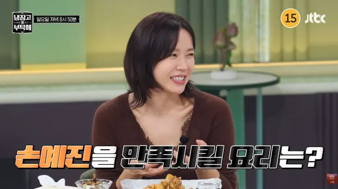 A smiling woman with short hair sitting at a table with food, discussing a recipe.