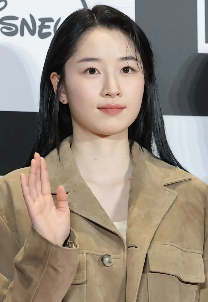 Won Ji-an with long dark hair wearing a beige jacket is waving, with a neutral expression on her face.