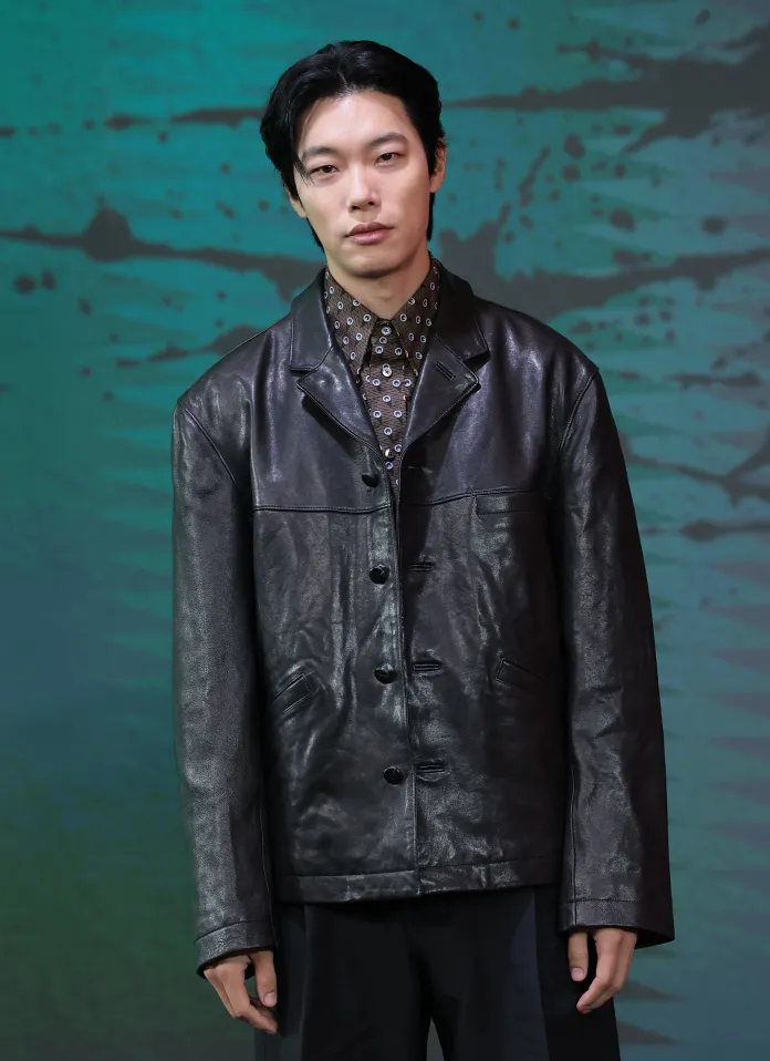 Ryu Jun-yeol wearing a black leather jacket and a patterned shirt stands confidently in front of an abstract teal background.