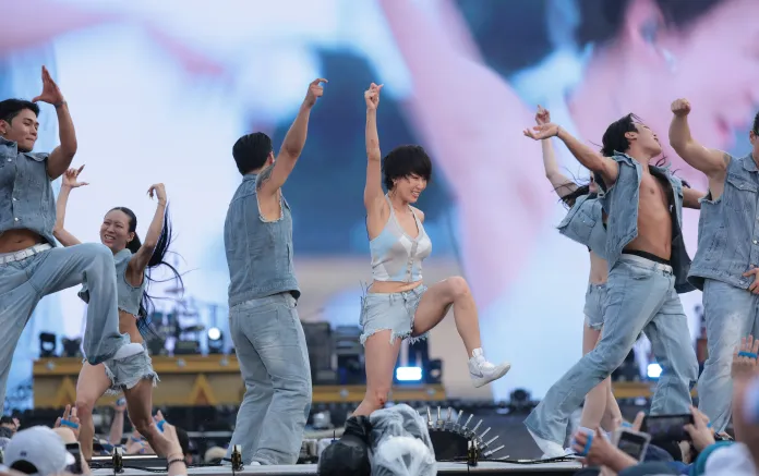 Hwasa featuring a group of six performers in denim outfits, showcasing energetic choreography on stage with vibrant lights in the background.