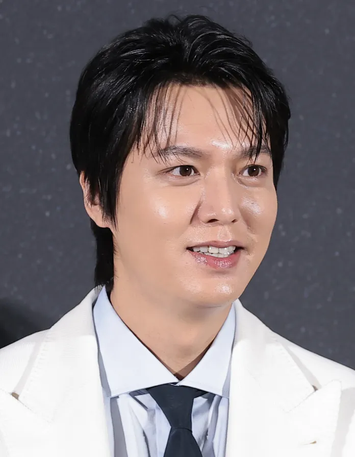 A man in a formal white suit and blue shirt, smiling, with dark hair and a neutral background.