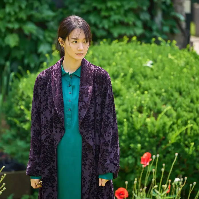 Shin Min-a standing in a garden, wearing a green shirt and a long purple coat, with green foliage and flowers in the background.