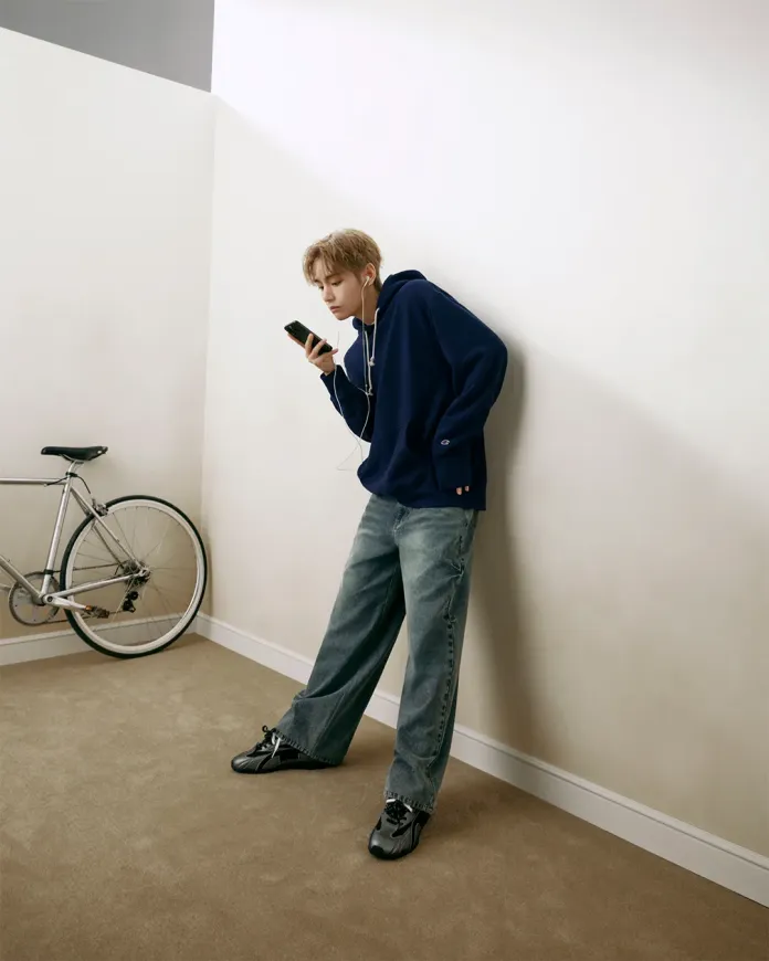 V in a navy hoodie and baggy jeans is leaning against a wall while holding a smartphone. They are wearing headphones and standing next to a bicycle in a minimalistic room.