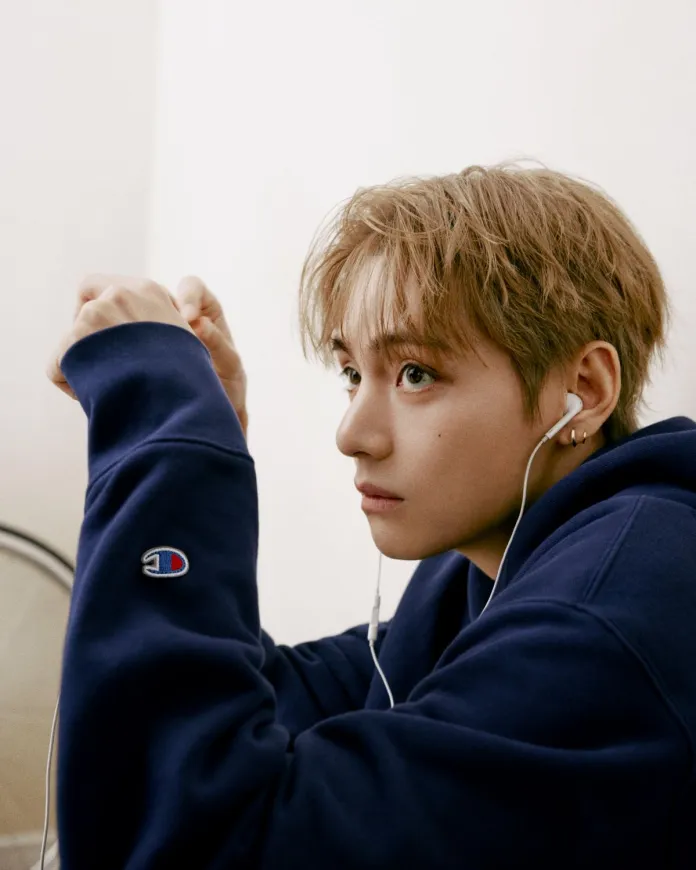 V wearing a navy blue hoodie with earphones, looking thoughtfully off to the side.