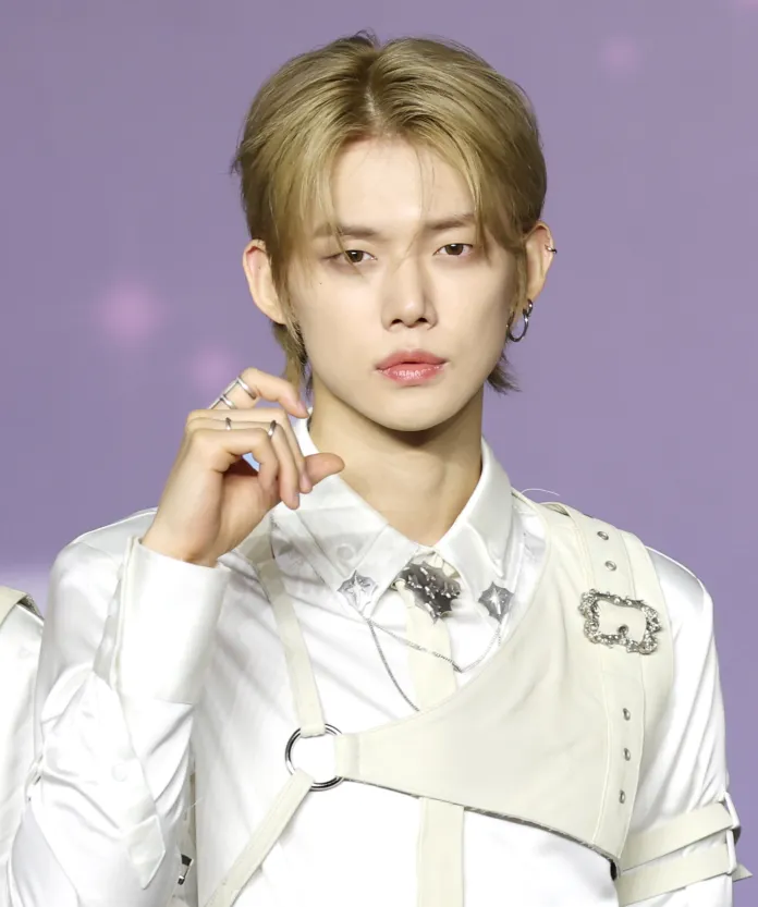 Yeonjun with styled blond hair, wearing a white shirt and a leather harness, poses confidently against a soft purple background.