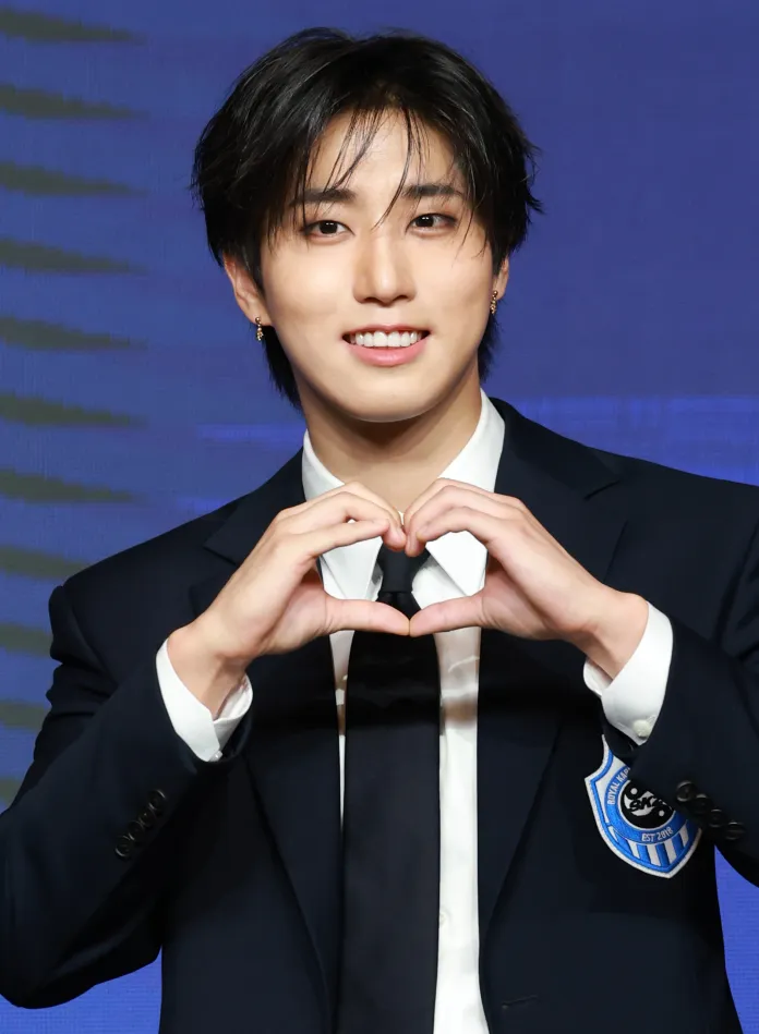 Stray Kids' Han in a dark suit and tie is making a heart shape with their hands, smiling at the camera.
