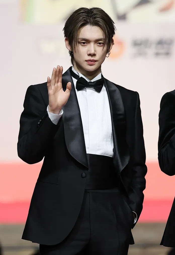 Yeonjun in a black tuxedo and bow tie waves at the camera, showing a confident expression.