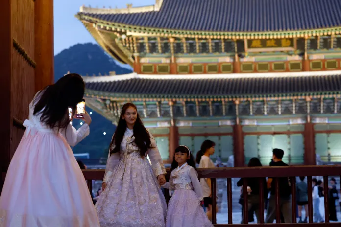 A woman in a traditional hanbok takes a photo of another woman and a young girl, both also in hanboks, in front of a brightly lit historical building during dusk.