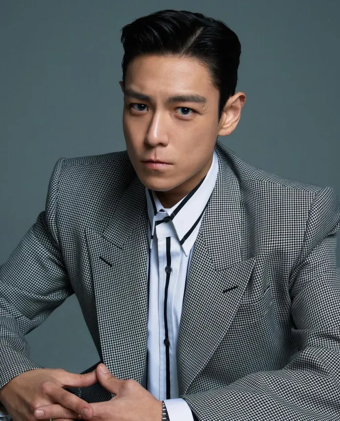 T.O.P wearing a stylish houndstooth patterned suit with a white shirt and black tie, sitting with a serious expression against a gray background.