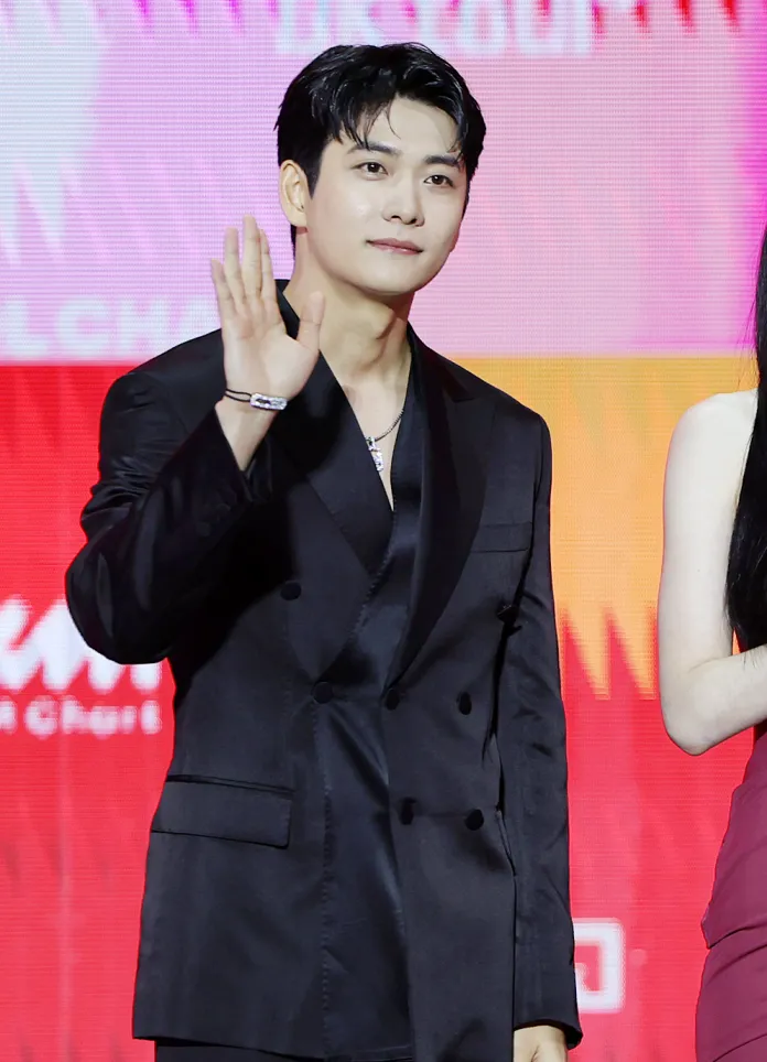 Kang Tae-oh in a black suit is smiling and waving at the camera, standing beside a woman in a red dress. The background features colorful graphics.
