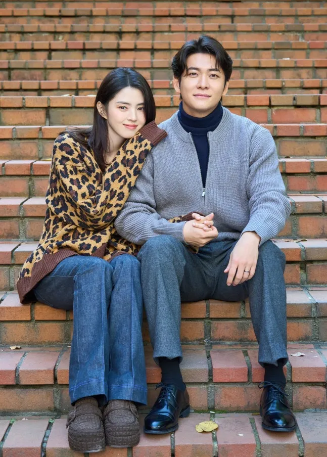 Han So-hee and Kang Tae-oh sitting on brick steps, Han So-hee wearing a leopard print sweater with long dark hair and Kang Tae-oh in a gray sweater. They are holding hands and smiling at the camera.