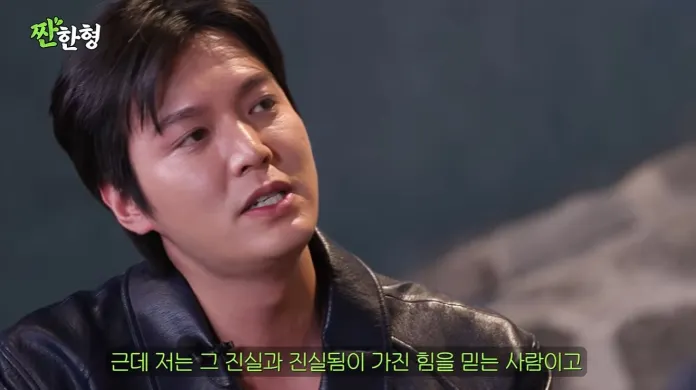 Lee Min-ho with short black hair wearing a leather jacket, speaking thoughtfully in a seated position, with a blurred background.