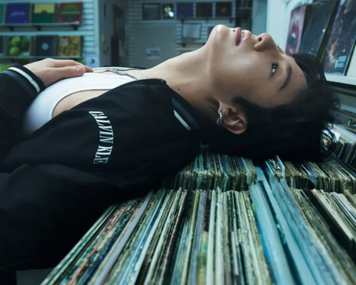 Calvin Klein lying on a stack of vinyl records, looking up. They are wearing a black jacket with a logo and have a piercing in their ear.