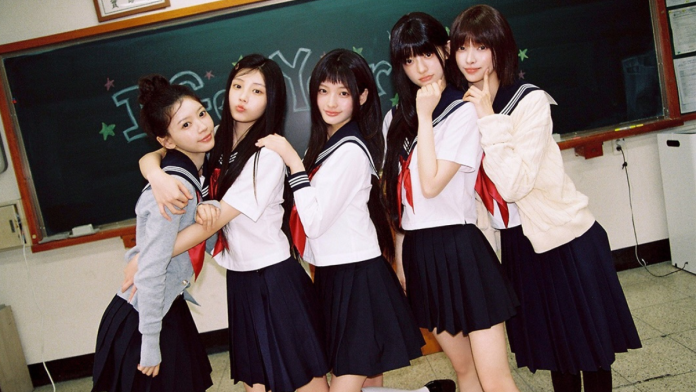 ILLIT members in school uniforms pose together in a classroom, with a chalkboard in the background. The first woman on the left is wearing a gray sweater and embracing another, while the others strike playful poses.