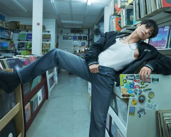 Calvin Klein posing in a vinyl record store, leaning against a shelf with a leg raised on a display. The person wears a black bomber jacket, a white tank top, and denim jeans, showcasing tattoos and accessories, while surrounded by colorful album covers and stickers.