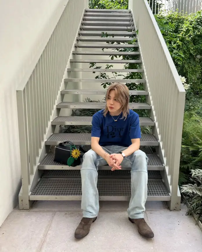 Jimin sitting on stairs surrounded by greenery, wearing a blue Dior t-shirt and light jeans, with a black bag and a sunflower.