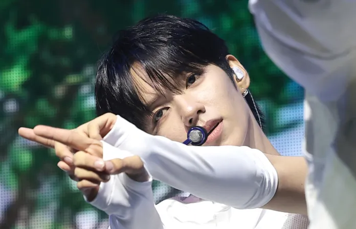 JL in a white outfit making a hand gesture on stage, with a microphone in their mouth and a focused expression.