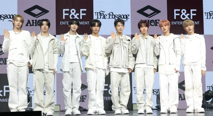 AHOF dressed in white and light-colored outfits, standing in a line with their hands raised in a greeting gesture. They are positioned in front of a branded backdrop featuring F&F Entertainment.