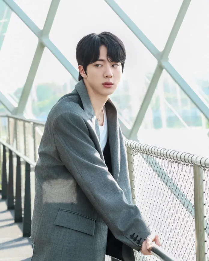 Jin in a gray blazer leans against a railing with a thoughtful expression, set against a backdrop of geometric glass architecture.