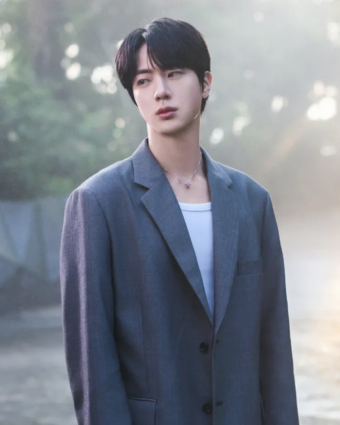 Jin wearing a gray oversized blazer over a white shirt, standing in a misty outdoor setting with a thoughtful expression.