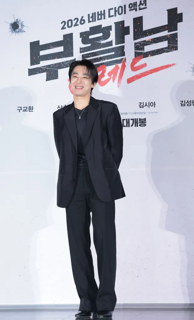 A smiling Koo Kyu-hwan in a black suit stands in front of a promotional backdrop for a movie titled '부활남' with the year 2026 indicated.