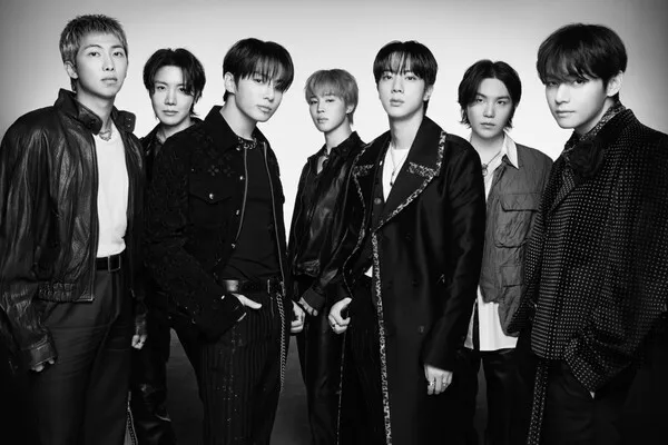 BTS standing in stylish outfits and posing confidently against a plain background.