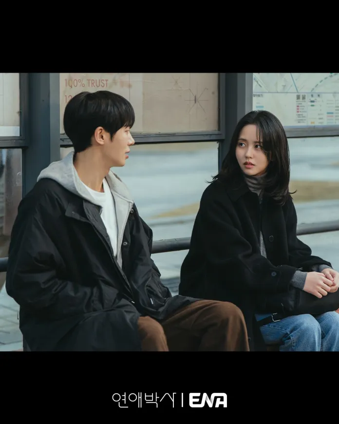 Choo Young Woo and Kim So Hyun sit on a bus stop bench, engaged in conversation. The man wears a black coat over a white shirt, while the woman is dressed in a black coat with a gray sweater. The background features a city scene.