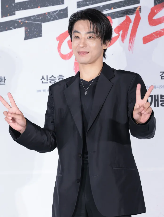 Koo Kyu-hwan in a black blazer and shirt poses with a peace sign at a promotional event.