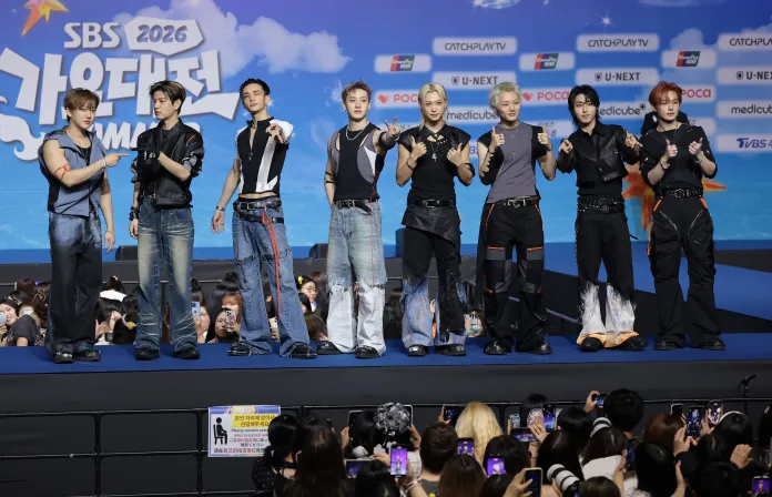 Stray Kids members on stage at an event, striking poses and smiling at the camera. They are dressed in stylish, fashionable outfits with unique designs. The background features a blue sky with clouds and event branding.