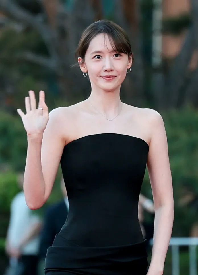 YoonA  in a strapless black dress waves while standing on a red carpet, with greenery in the background.