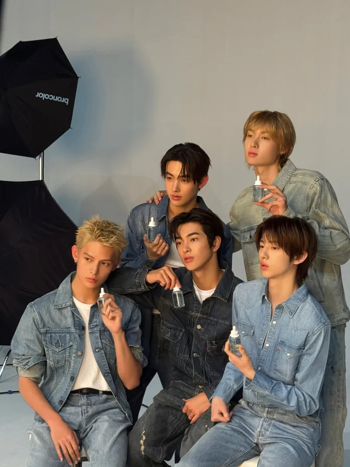 CORTIS members in denim outfits pose together, each holding a bottle. The background features studio lighting equipment, creating a professional photographer's setting.