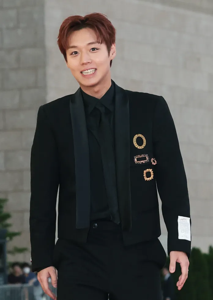 A smiling Park Ji-hoon in a black suit with a black tie, walking on a red carpet at an event.
