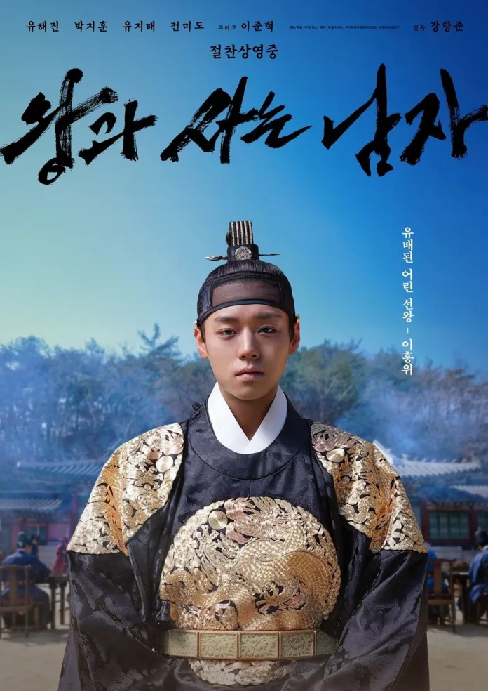 Park Ji-hoon dressed in traditional royal attire, featuring intricate gold embroidery, a black hat, and a serious expression, standing outdoors with a historical backdrop.