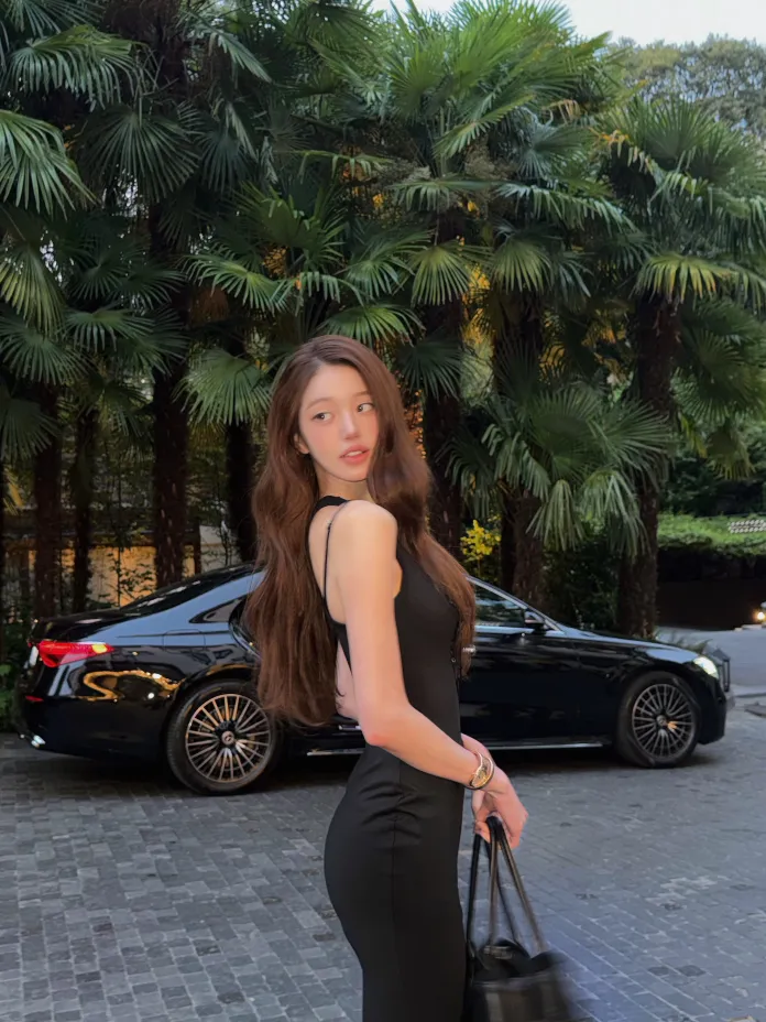 Jang Wonyoung wearing a black dress stands outdoors with palm trees in the background. A luxury car is parked nearby.