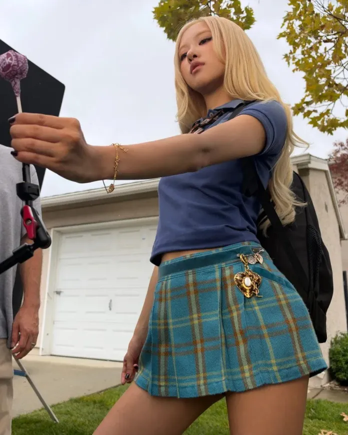 ROSÉ with long blonde hair poses outdoors, wearing a blue polo shirt and a plaid mini skirt. She holds a lollipop in one hand while a backpack hangs from her shoulder.