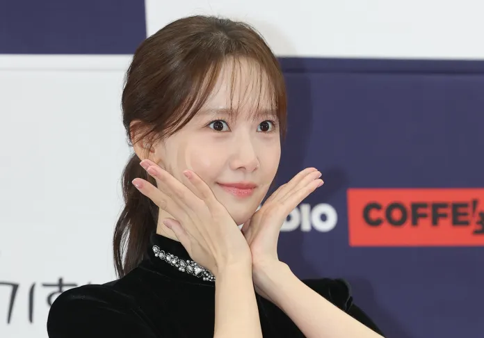 YoonA with long hair smiles and poses with her hands gently under her chin, wearing a black outfit.