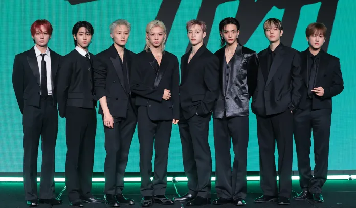 Stray Kids members dressed in black suits, posing together for a promotional event against a green backdrop.