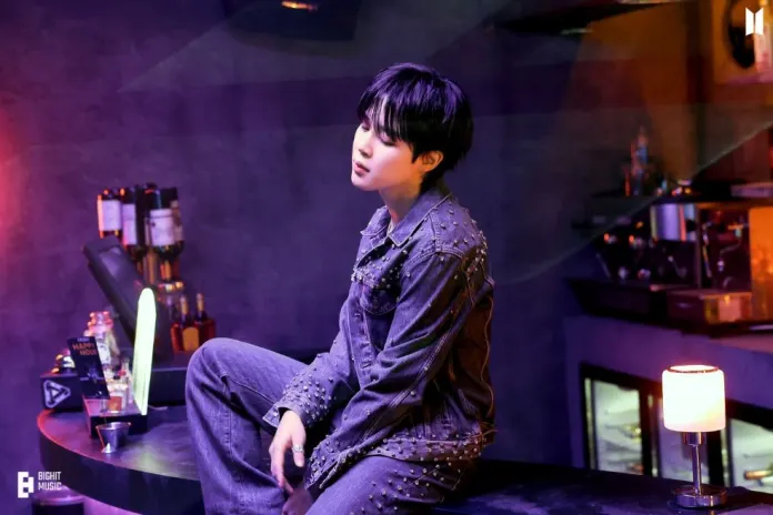 Jimin with short black hair seated elegantly on a bar, wearing a denim outfit with studs, in a dimly lit, stylish bar environment.