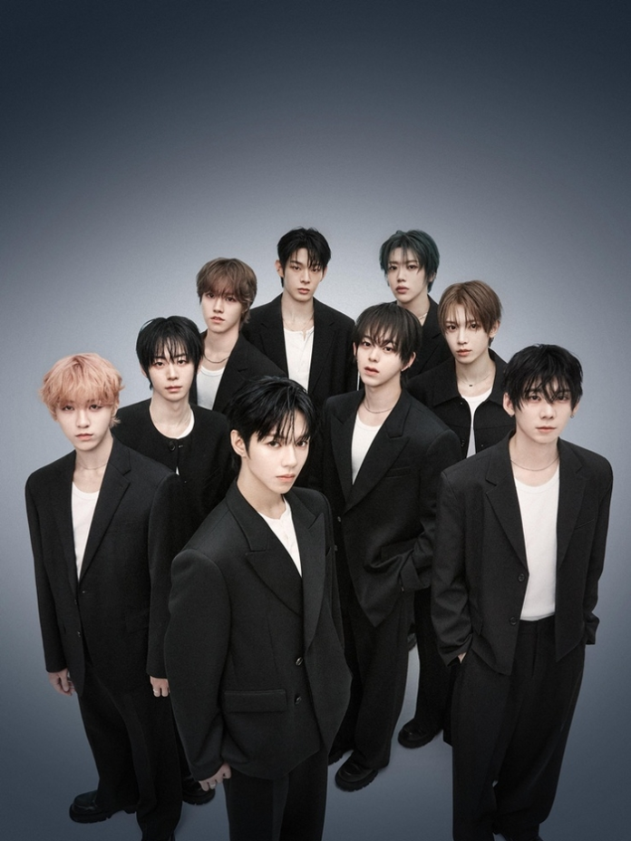 A group of ten young men dressed in black suits and white shirts, posing together in a stylized, dramatic setting with a gradient background.