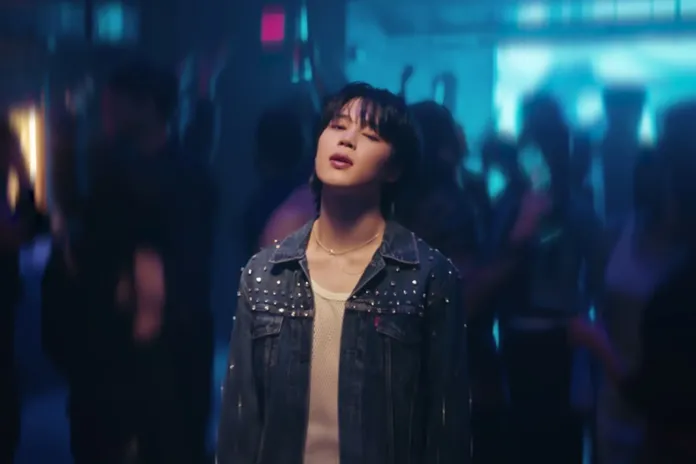Jimin wearing a denim jacket with studs stands in a dimly lit party atmosphere, surrounded by blurred figures dancing and enjoying the music.