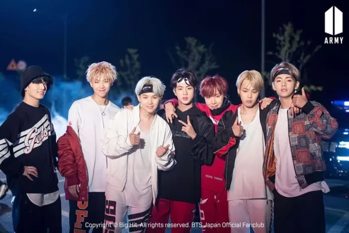 BTS posing together outdoors at night, with a subtle smoky background. They are dressed in casual streetwear, making various hand gestures and smiling at the camera.