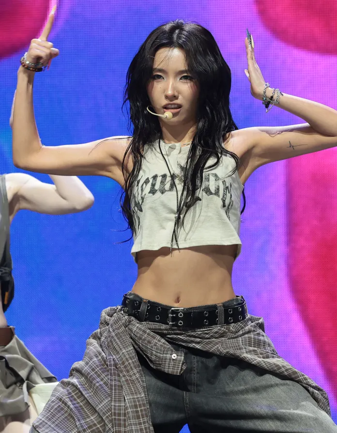 Soyeon with long, dark hair strikes a dynamic pose on stage, wearing a cropped tank top and wide-leg jeans, with a microphone in their mouth and colorful stage lighting in the background.