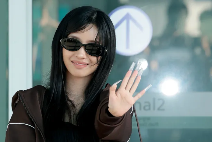 Soyeon with long black hair and sunglasses waves at the camera, dressed in a brown jacket, with distinctive long nails. The background shows an airport sign.