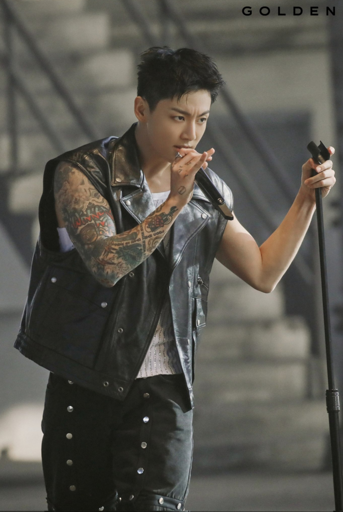 Jungkook performing on stage, wearing a black leather vest and showcasing arm tattoos, with a focused expression while holding a microphone.