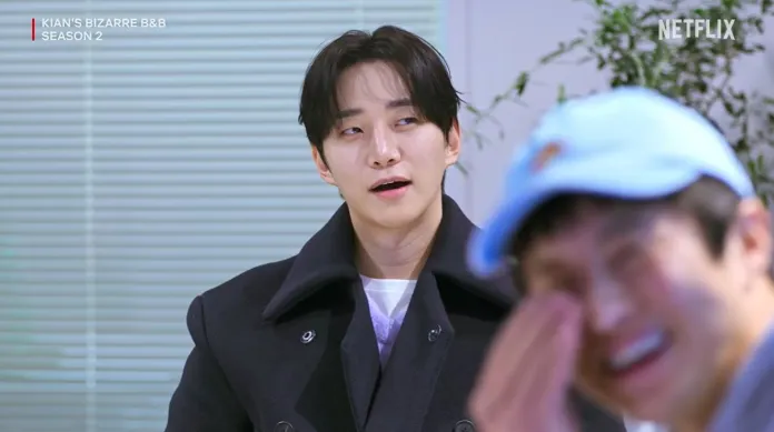 Lee Junho in a black coat sits in front of a blurred background, looking surprised or amused, while another man in a blue cap smiles and covers his face, suggesting a lighthearted moment.