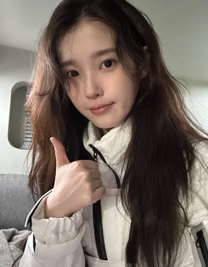 IU with long hair wearing a white jacket gives a thumbs up, smiling at the camera indoors.
