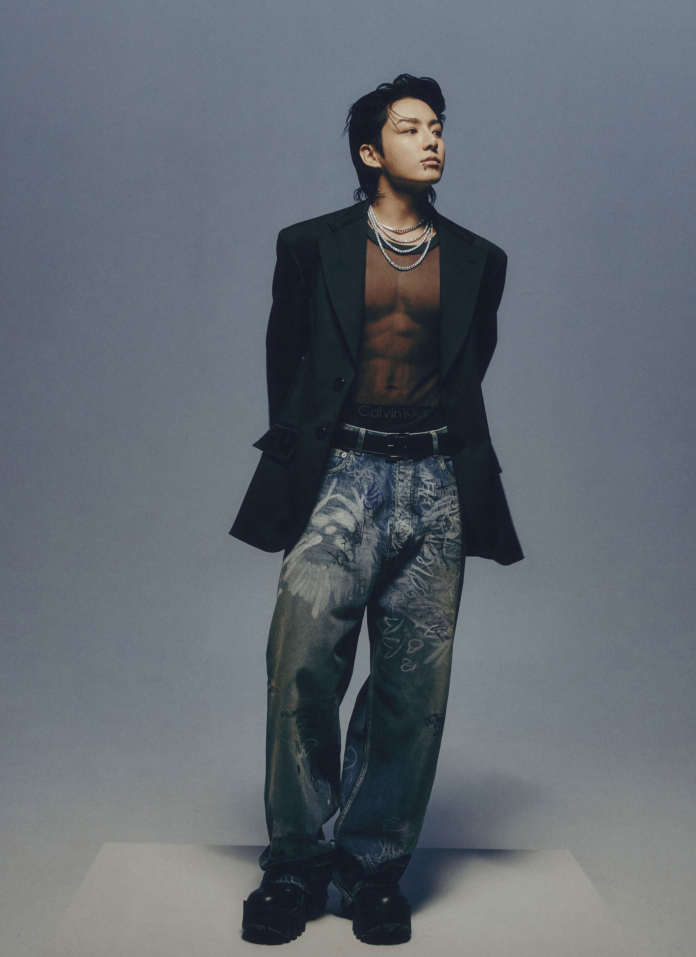 Jungkook striking a pose in a stylish outfit featuring a sheer black top and large oversized blazer, paired with distressed jeans and chunky black shoes against a gray background.
