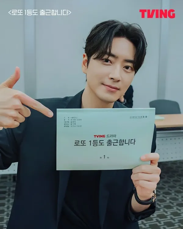 Lee Joon-hyuk in a black jacket points to a document that reads 'TVING 드라마 로또 1등도 출근합니다' while smiling, in a room with a table and chairs.