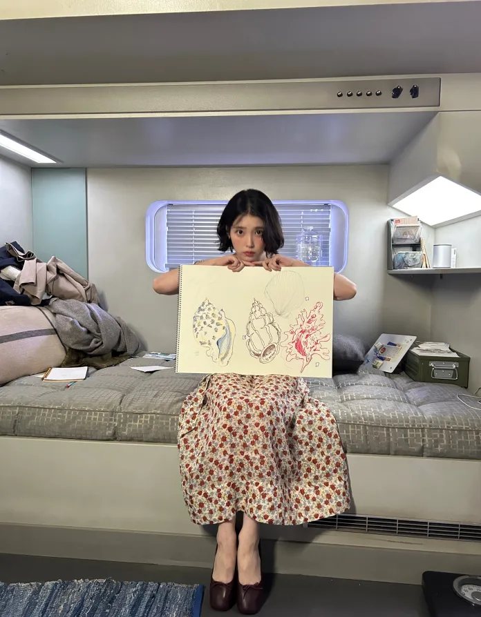 IU in a floral dress holding a sketchbook with drawings of shells and sea creatures, sitting on a bed in a cozy interior space.