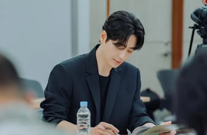 Lee Joon-hyuk in a black blazer sits at a table, attentively writing in a notebook, with a water bottle placed nearby.
